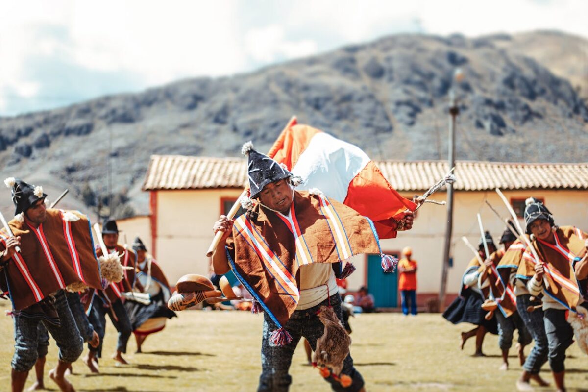 Men in traditional attire perform a cultural dance with flags at an outdoor festival in the mountains.